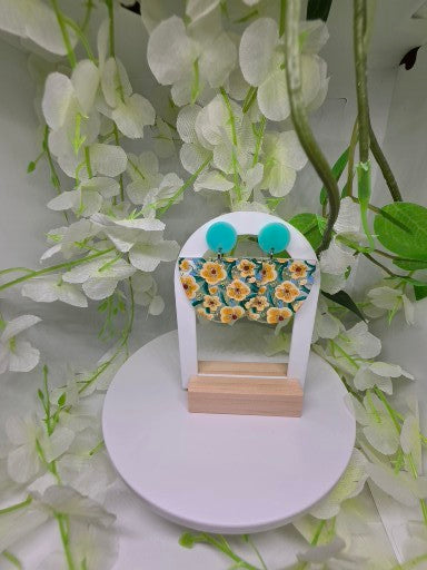 A pair of handmade fabric earrings displayed on a small wooden stand, set against a background of white artificial flowers. The earrings feature a teal and yellow floral pattern with two bright turquoise blue circular studs.
